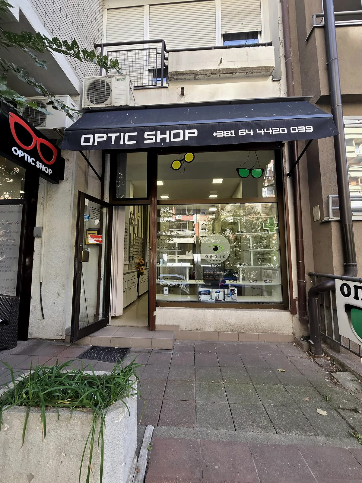 Ulaz u Optic Shop: crna tenda sa natpisom i brojem telefona, levo tabla sa crvenim naočarima